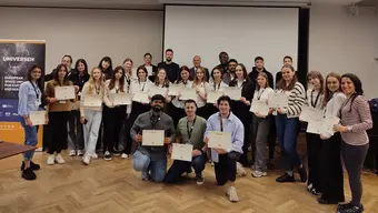 Grupa młodych ludzi pozuje do zdjęcia w sali konferencyjnej, trzymając certyfikaty. W tle baner z napisem 'UNIVERSEH' oraz ekran projekcyjny