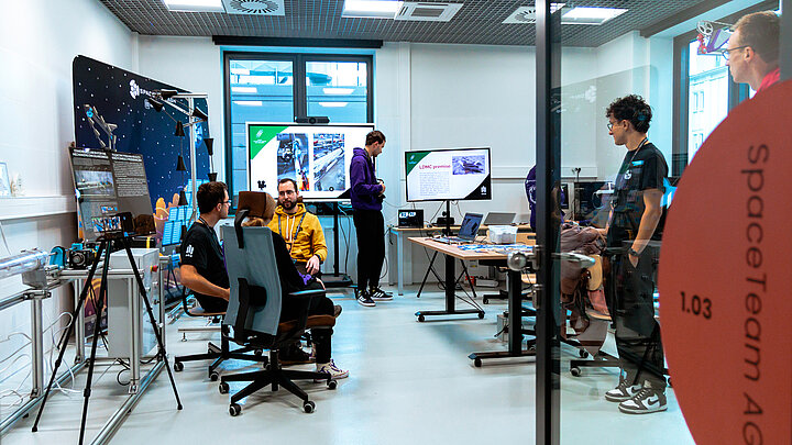 GRupa młodych ludzi w laboratorium. W tle dwa duże ekrany oraz kilka laptopów na stołach. Po lewej proptotyp urządządzenia do transportu sypkich substancji. 